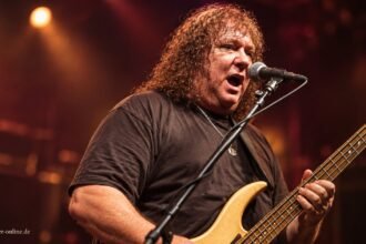 Steve Priest mit Bass und Mikrofon vor einem gestalteten Bühnenhintergrund
