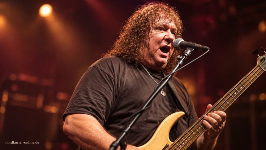 Steve Priest mit Bass und Mikrofon vor einem gestalteten Bühnenhintergrund